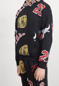 Sudadera con capucha negra con gráficos vibrantes, incluyendo anillos de campeonato dorados, motivos de toros y números rojos. Cuenta con capucha a rayas y bolsillo frontal.