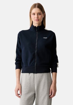 Blonde Frau mit einer schwarzen Reißverschlussjacke mit weißen Streifen an den Ärmeln und dem "GUESS"-Logo auf der Brust, kombiniert mit hellgrauen Hosen.