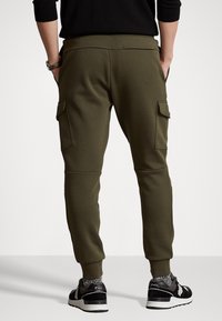 Olive green joggers with elastic cuffs, side cargo pockets, and a smooth fabric texture. Worn with black sneakers and gray socks.