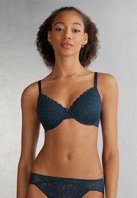 Marintrådfärgad spetsbehå med vågiga kanter och tunna remmar, med blommönster och en liten rosett i mitten.