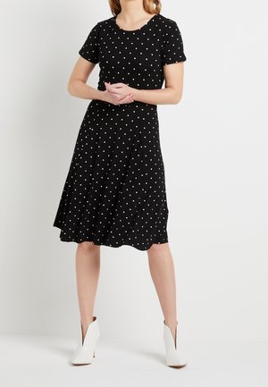 Femme portant une robe noire mi-longue à pois blancs et des bottines blanches, debout devant un fond clair uni.