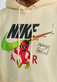 Sweatshirt creme com gráfico frontal apresentando o texto "NIKE", um pimento cartoon com óculos de sol, um swoosh verde e ténis amarelos. Duas cordas de ajuste.