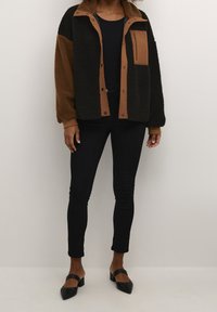 Musta ja ruskea fleece-takki, jossa on ruskean sävyinen nappikiinnitys ja vetoketjullinen tasku. Yhdistetty mustiin, istuviin farkkuihin ja mustiin, teräväkärkisiin mules-kenkiin.