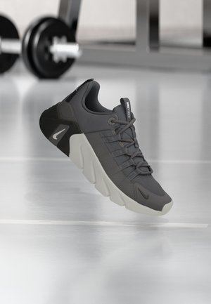 Tmavosivá a čierna športová obuv Nike Free vznášajúca sa nad lesklou podlahou fitnescentra s rozmazanými jednoručnými činkami v pozadí.