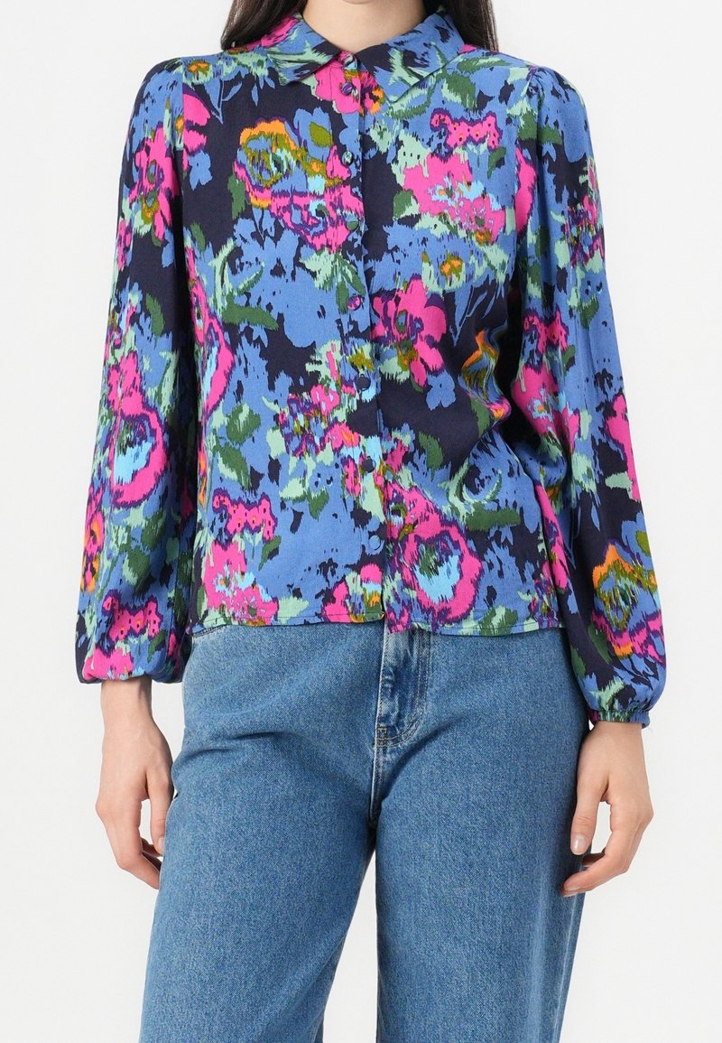 Blouse florale dans des teintes de bleu, rose et vert, avec un devant à boutons, un col et des manches longues avec des poignets froncés.