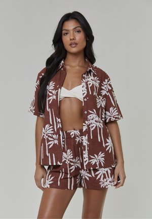 Kobieta ubrana w brązową koszulę na krótki rękaw na guziki oraz pasujące do niej szorty z białym nadrukiem palm, zestawione z białym krótkim topem.
