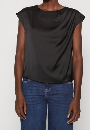 Vrouw draag een mouwloze zwarte satijnen blouse, gedeeltelijk ingestopt in een blauwe spijkerbroek met hoge taille, staand met ontspannen armen langs haar zij.