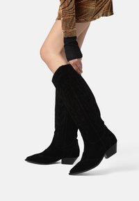 Knee-high crne ante čizme u stilu kaubojaca s šiljastim nosom, teksturiranim šavovima i kutnim izrezima duž osovine. Blok potplati upotpunjuju dizajn.