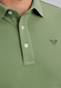 Zelena polo majica, teksturiran materijal, tri dugmeta, izvezen logo u tamnijoj nijansi, suptilna kragna i glatka završna obrada.