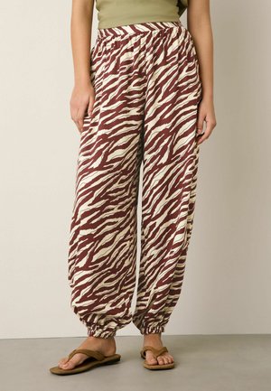 Femme debout portant un pantalon ample avec des chevilles élastiques imprimé zèbre marron et crème, un haut sans manches vert olive, et des sandales plates marron.