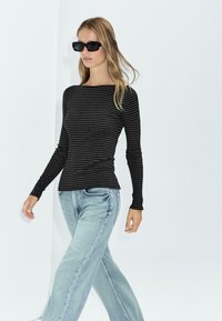 Zwart en grijs gestreept oversized shirt met een bootnek, gecombineerd met lichtblauwe wijde spijkerbroek. Het model draagt donkere zonnebril.