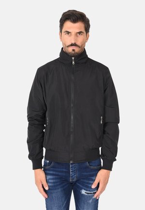 Uomo che indossa una giacca nera con zip e collo alto con polsini a coste, abbinata a jeans blu strappati, in piedi davanti a uno sfondo bianco semplice.