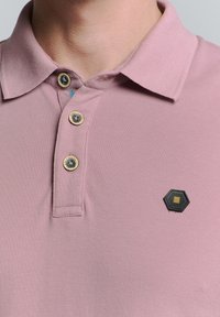 Polo shirt in lichtroze stof met een gestructureerde afwerking, voorzien van drie houten knopen en een hexagonaal zwart logo op de borst.