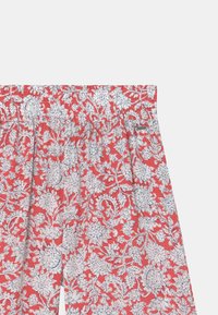 Rode elastische taillebroek met witte bloemen- en loofpatronen en een klein rechthoekig metalen merkteken aan de rechterkant.