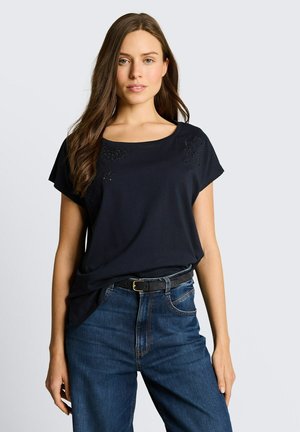 Kobieta z długimi brązowymi włosami, ubrana w czarną bluzkę z krótkim rękawem i niebieskie jeansy z czarnym paskiem, stojąca na jasnym, jednolitym tle.