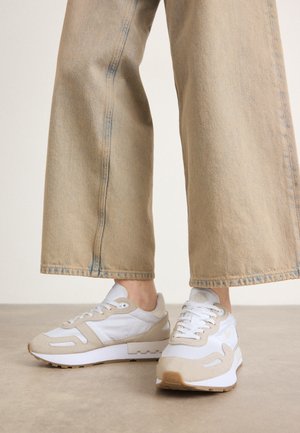 Beige wijde broek gecombineerd met witte en beige casual sneakers op een neutrale vloerachtergrond.