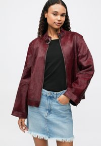Veste en cuir bordeaux avec col montant, fermeture éclair à l'avant et manches larges, associée à un haut noir et une jupe en denim déchirée bleu clair.