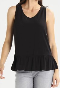 Top negro sin mangas con escote en V y dobladillo con volantes, combinado con jeans gris claro, mostrado de los hombros a las caderas.
