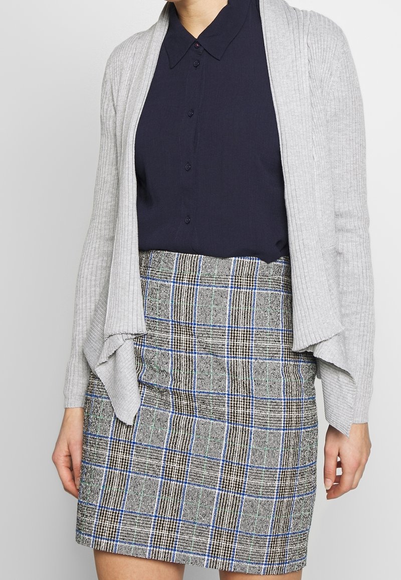 Femme portant une chemise noire à boutons, un cardigan gris clair ouvert sur le devant et une jupe à carreaux longueur genou avec des lignes bleues et noires.