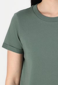 Grön bomull t-shirt med rund hals och korta ärmar. Ärmarna är upprullade. Tyget verkar slätt med en subtil textur.