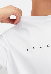 Witte katoenen t-shirt met een ronde halslijn, met aan de voorkant een zwart geprinte tekstlogo "JACK", vastgehouden bij de kraag door een hand.