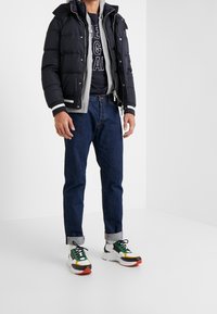 Persona vistiendo una chaqueta de plumas negra sobre una sudadera gris y jeans azul marino, combinada con zapatillas blancas, verdes, amarillas y rojas, de pie en un suelo blanco.