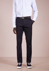 Zwarte op maat gemaakte broek met een gladde textuur, voorzien van zijzakken en riemlussen, gecombineerd met zwarte sneakers met witte veters.