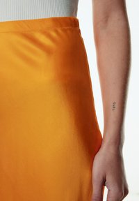 Oranje satijnen rok met een soepele textuur en een hoge tailleontwerp. Zichtbare arm met een tatoeage die de zijkant van de rok vasthoudt.