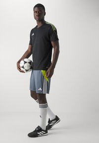 Svart kortermet trøye med grønne detaljer, grå shorts med sideløsning, hvite sokker med svarte striper, og svarte fotballsko.