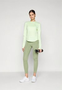 Langärmeliges, hellgrünes Sportoberteil, eng anliegende olivgrüne Leggings und weiße Sportschuhe. Model hält eine schwarze Wasserflasche. Durchgehend glatte Textur.