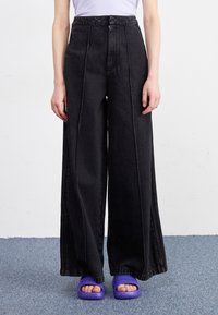 Široke črne jeans hlače imajo visok pas, sprednje zadrgo in podvojene šive. Usklajene so s purpurnimi natikači.