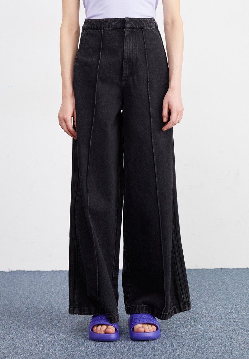 Široke črne jeans hlače imajo visok pas, sprednje zadrgo in podvojene šive. Usklajene so s purpurnimi natikači.
