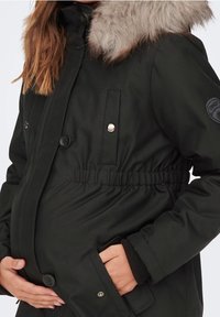 Svart gravidjacka med huva i konstpäls, elastisk midja, två framfickor och tryckknappar. Mjuk textur och varm design.