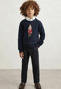 Jūrinės mėlynos spalvos megztinis su šokėjo dizainu centre, derinamas su tamsiomis kelnėmis ir odiniais loafer'iais. Audinys yra minkštas.