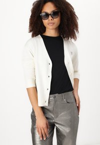 Cardigan tricoté blanc avec un décolleté en V et six boutons noirs, associé à un haut noir et un pantalon argent métallique. Le modèle porte des lunettes de soleil noires.