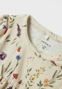 Tissu beige clair comportant divers motifs floraux dans des couleurs comme le violet, le rouge, le bleu et le jaune ; détails de décolleté scallopé.