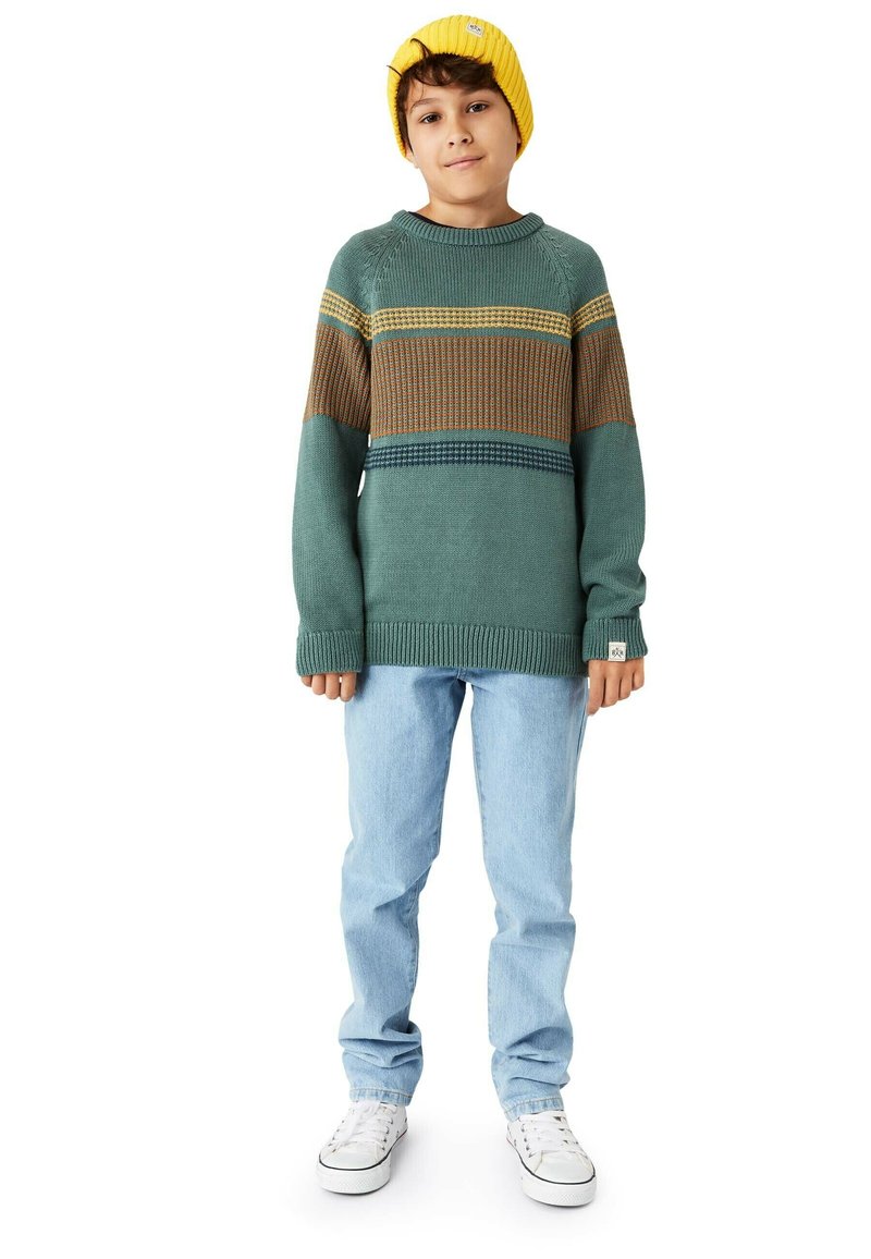 Stickad tröja i teal med gula, bruna och blå horisontella ränder; rund hals, nedhasade axlar, matchad med ljusblå jeans och vita sneakers.