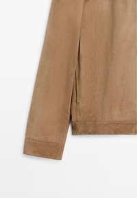 Beiger Wildleder-Jackenärmel mit sichtbaren Nähten und einem vertikalen Nahtdetail nahe der Manschette auf weißem Hintergrund.