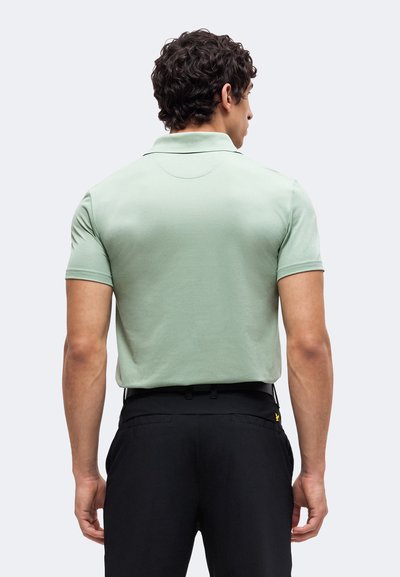 Homme de dos portant un polo vert clair à manches courtes rentré dans un pantalon noir sur un fond uni.