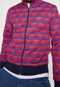 Veste à fermeture éclair rouge et bleue avec un motif géométrique 3D, poignets et ourlet côtelés, avec un petit logo vert sur la poitrine.