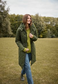 Mujer sonriendo y caminando en un abrigo acolchado verde, suéter verde, jeans azules y botas grises en un campo de hierba con árboles de fondo.