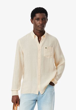 Hombre con camisa de botones color crema con un bolsillo en el pecho y jeans azul claro, sosteniendo la correa de una bolsa marrón claro con una mano.