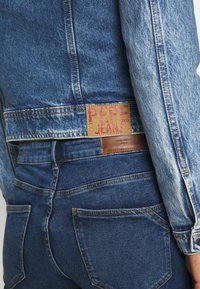 Jeanskappa med ljusblå ärmar och mörkblå kropp; har en "PEPE JEANS"-etikett i rosa stickning, tillsammans med mörkblå jeans.