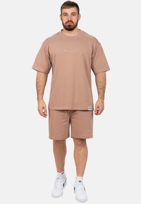 T-shirt ample en coton marron à manches courtes avec logo brodé, accompagné d'un short assorti. Chaussures de sport blanches complètent la tenue.