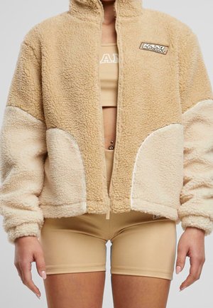 Beige und cremefarbene Sherpa-Jacke mit Reißverschluss, getragen über einem hellbraunen Sport-BH und passenden hochgeschnittenen Biker-Shorts, sichtbare Bauchnabelpiercings.