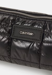 Czarna pikowana nylonowa saszetka z srebrnym suwakiem. Posiada czarną skórzaną metkę z wytłoczonym napisem "Calvin Klein". Gładka i błyszcząca powierzchnia.