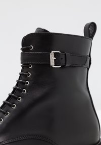 Botín negro de cuero hasta el tobillo con ojales metálicos, un pequeño detalle de hebilla plateada y textura suave, con un diseño de punta redondeada.