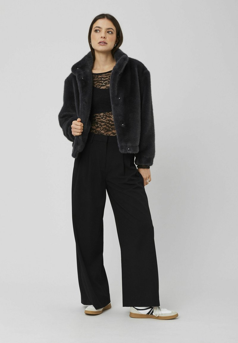 Veste en fausse fourrure gris foncé, avec un col châle, des détails en boutons et une coupe décontractée, portée avec un pantalon large noir et des chaussures blanches.