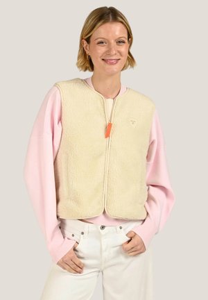 Femme souriante portant une veste polaire crème zippée sur un pull rose clair avec un pantalon blanc, posant les mains dans les poches.