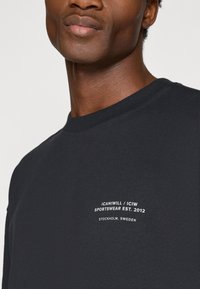Melna kokvilnas sporta krekls ar apaļu kaklu, ar baltu tekstu uz kreisā krūšu sāna: "ICANIWILL / ICIW SPORTSWEAR EST. 2012, STOCKHOLM, SWEDEN."